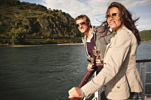 Two people stand on the sun deck of a river cruise ship, enjoying the view of the Rhine with a glass of white wine, surrounded by lush hills and blue skies.