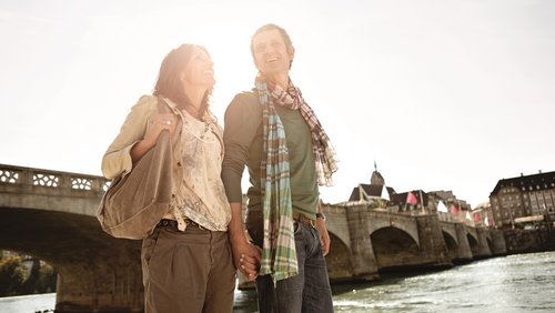 A couple is walking along the banks of the Rhine in Basel in the sunshine. In the background, there is a stone bridge that crosses the river.