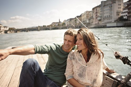 A couple is sitting and relaxing on a boat on the Rhine in Basel, enjoying the view in the sunshine. In the background, there are buildings on the shore and a bridge.