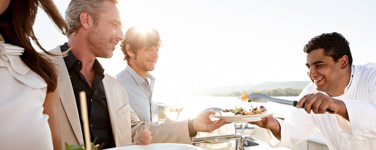 Three people are standing at the barbecue on the sun deck of a river cruise ship, and the cook is serving them food from the grill, with the river and trees in the background.