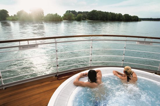 Two people are relaxing in a hot tub on the deck of a river cruise ship with a view of the Rhine in the sunshine.