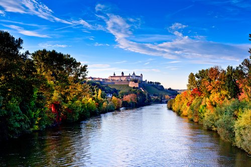Fluss mit bunten Herbstbäumen an beiden Ufern und Festung Marienberg auf einem Hügel in Würzburg unter blauem Himmel