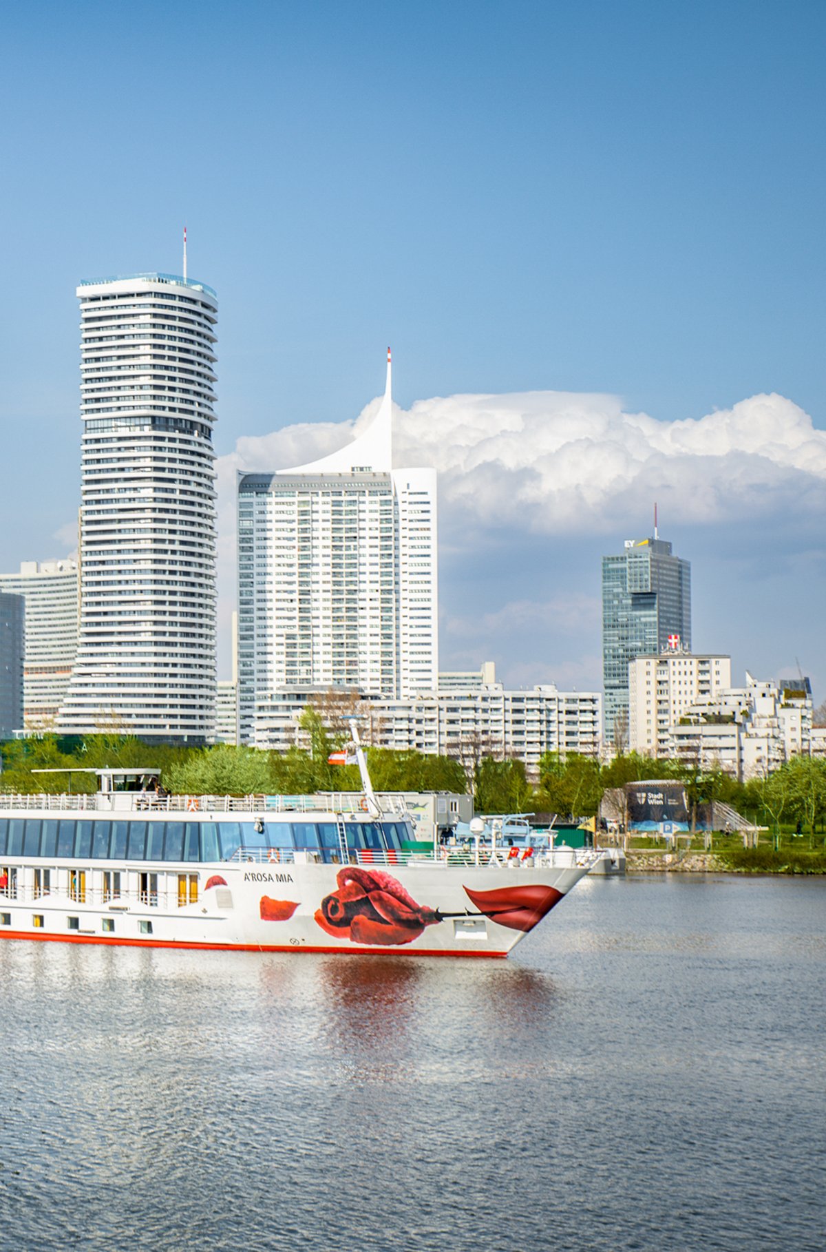 Die A-ROSA MIA fährt in Wien auf der Donau vor moderner Skyline mit Donauturm, Hochhäusern und Uferpromenade bei Tageslicht.