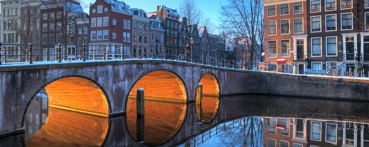 Brücke mit drei Bögen über einen Kanal in Amsterdam, beleuchtet von warmem Licht, mit historischen Häusern und klarer Spiegelung im Wasser