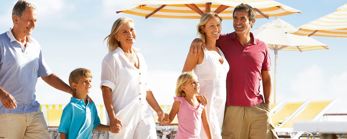 Three generations of a family are walking and laughing on a sun deck with yellow and white umbrellas and deck chairs.