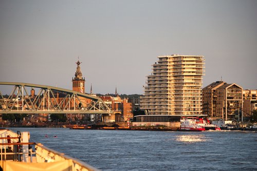 View of the riverside promenade of Nijmegen with a modern tall residential building, a historic church spire, and a bridge over the river.