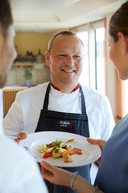 A cook in a white jacket and striped apron serves guests a plate with a fine, artfully arranged dish.