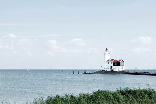 A lighthouse on the IJsselmeer with reeds in the foreground and sailboats on the sea, under a cloudy sky.