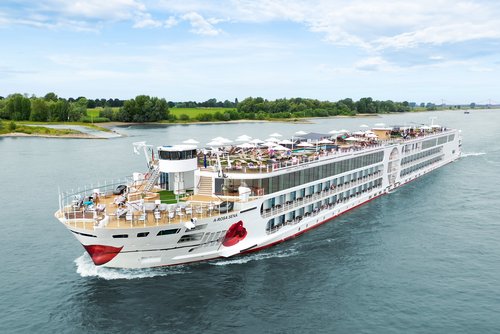 The A-ROSA SENA sails along the Rhine, the area with deck chairs and sunshades is well attended, with the riverbank featuring green landscapes and trees in the background.