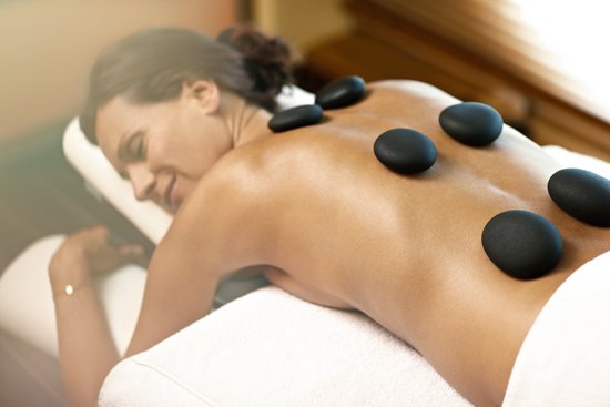 A woman lies relaxed and smiling on a massage table, while hot stones are placed on her back, surrounded by a tranquil spa atmosphere.