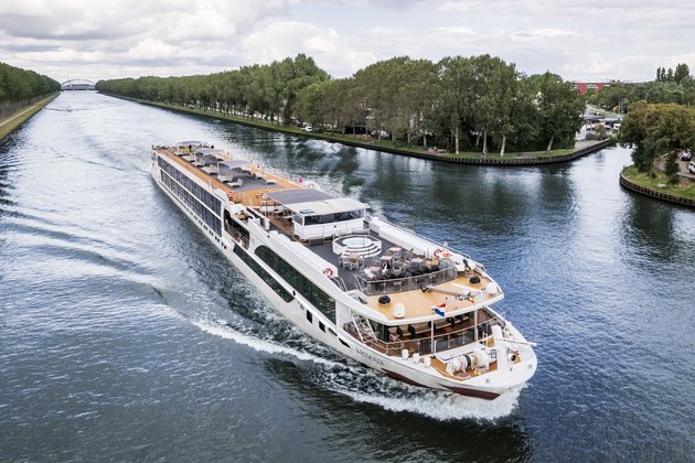 The A-ROSA ALEA is sailing along an urban shore with trees, with a bridge in the background. The sun deck is equipped with a whirlpool, sun loungers, and parasols.