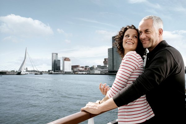A couple leans against the railing of a river cruise ship, looking at the Rhine and the skyline of Rotterdam.