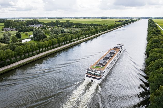 A river cruise ship sails on the Rhine past a small village, green fields, and trees on the shore. The sun deck is equipped with a whirlpool, sun loungers, and parasols.