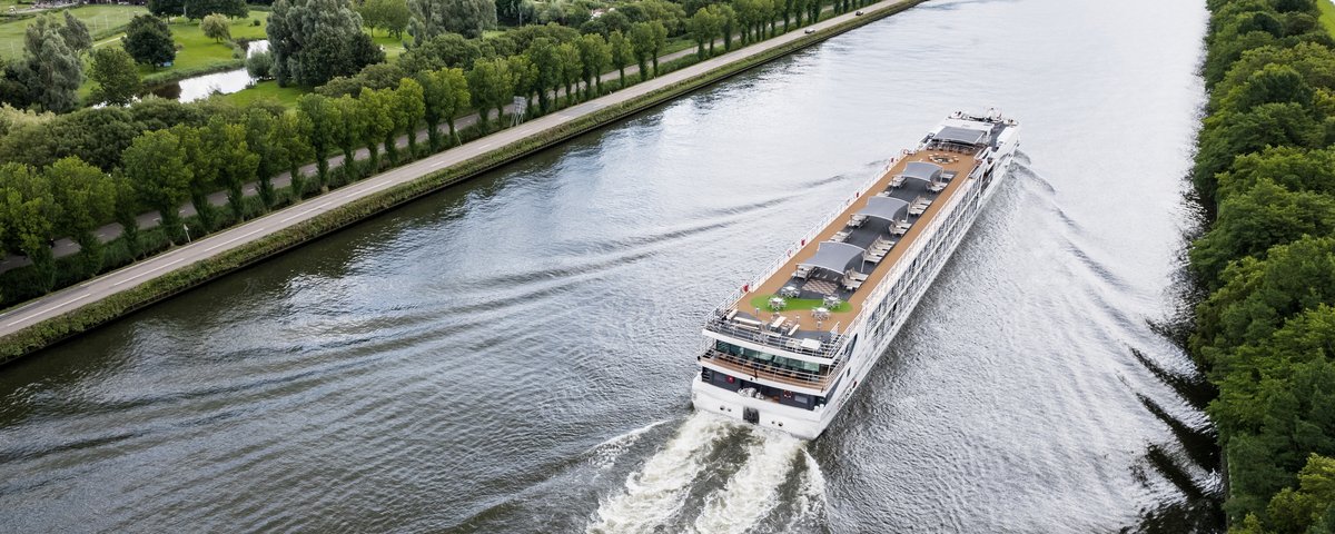 A river cruise ship sails on the Rhine past a small village, green fields, and trees on the shore. The sun deck is equipped with a whirlpool, sun loungers, and parasols.