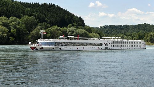 In the afternoon, the A-ROSA SILVA sails along the Rhine past green, wooded hills on the shore; the sky is blue and slightly cloudy.