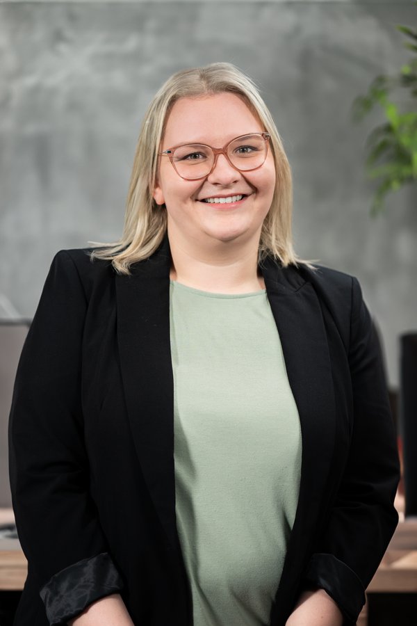 Elisa Gunert smiling with blonde hair, glasses, green shirt, and black blazer against a gray background