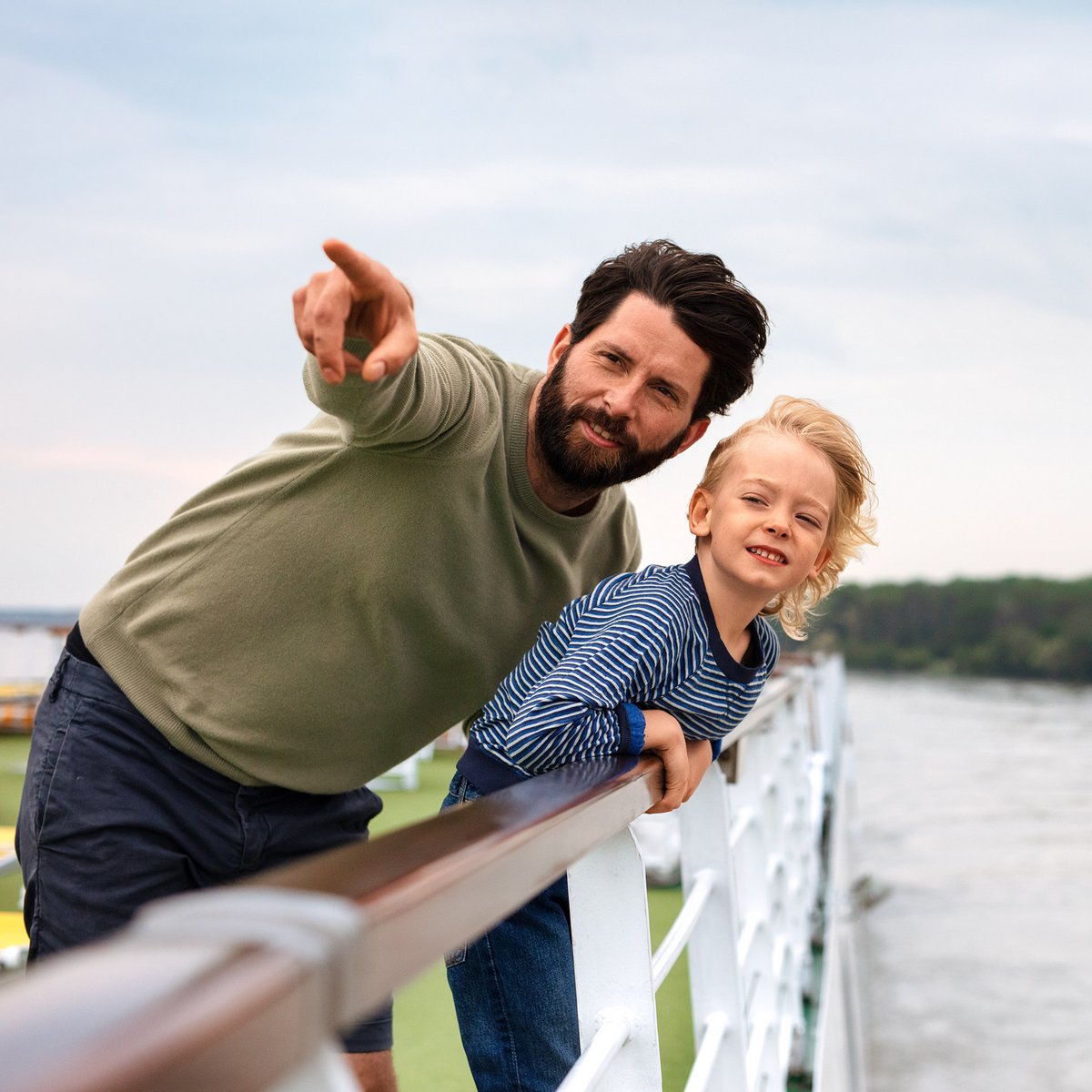 Father and son stand on the deck of a river cruise ship and look over the railing at the river. The father points to something in the distance.