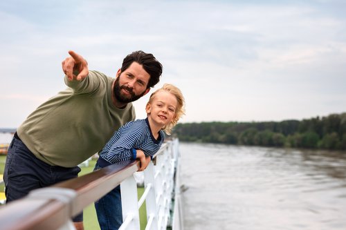 Father and son stand on the deck of a river cruise ship and look over the railing at the river. The father points to something in the distance.