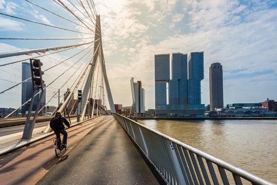 Cyclist on the Erasmus Bridge in Rotterdam with a view of modern high-rise buildings on the riverbank under a cloudy sky