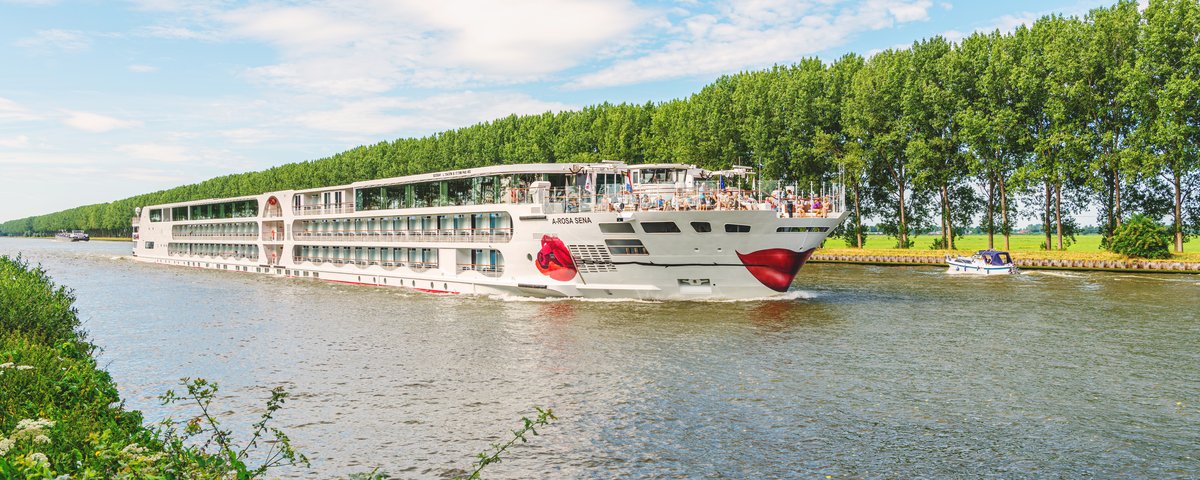The A-ROSA SENA sails through a Rhine canal past a row of trees. The sky is blue and slightly cloudy.