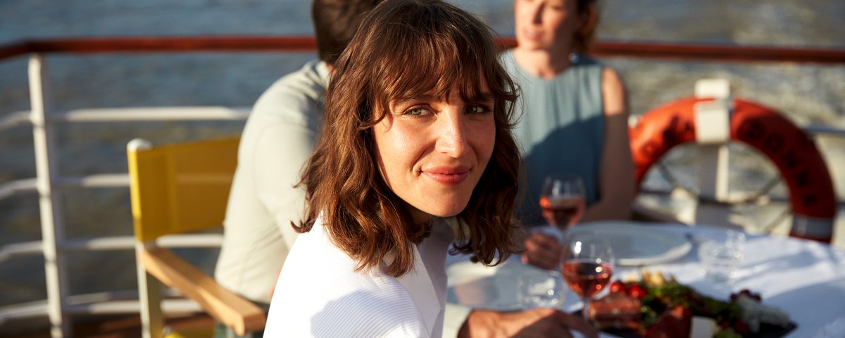 A woman with brown hair and a white sweater smiles on a boat, with two other people and a set table with wine and snacks in the background.
