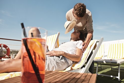 A woman is lying on a sun lounger on the deck of a river cruise ship. A man is standing behind her, providing her with shade using a hat. In the foreground, there is a drink with an orange slice.