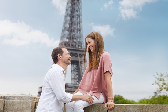 Couple in front of the Eiffel Tower, man in a white shirt holding a woman in a pink top sitting on a wall.