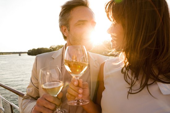 A couple stands at sunset with a glass of wine in hand at the railing of a riverboat. In the background, the river and a riverside landscape.