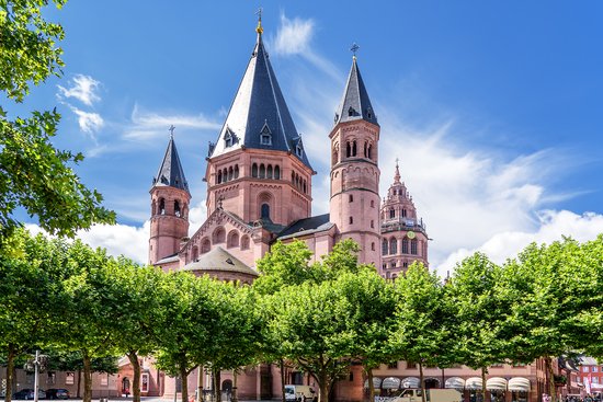 The Mainz Cathedral St. Martin with its distinctive red sandstone towers and pointed roofs, surrounded by green trees and a blooming front garden under a blue sky