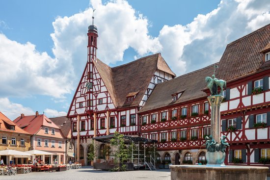 Historic half-timbered houses on a market square in Forchheim, featuring a fountain and a column topped with a statue of a horseman, under a slightly cloudy sky.