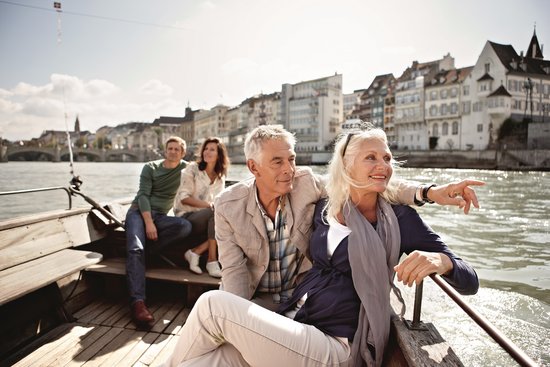 Two couples are sitting and relaxing on a boat on the Rhine in Basel, enjoying the view in the sunshine. In the background, there are buildings on the shore and a bridge.
