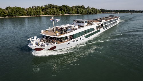 A river cruise ship is sailing along a wooded bank on the Rhine. The sun deck is equipped with a whirlpool, sun loungers, and sun sails.
