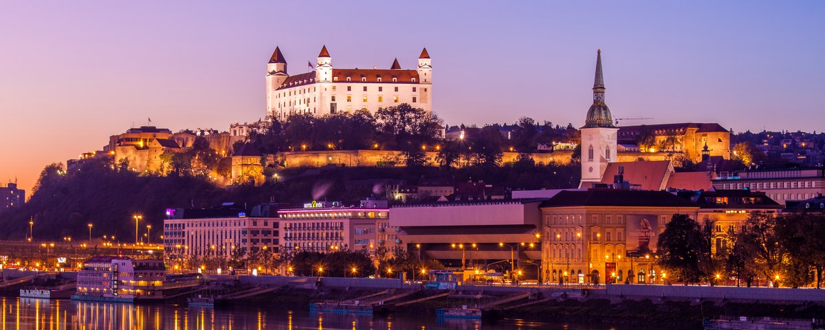 View of the Danube riverbank in Bratislava at sunset with Bratislava Castle in the background, surrounded by trees and the city. In the foreground, the lights are reflected in the river.