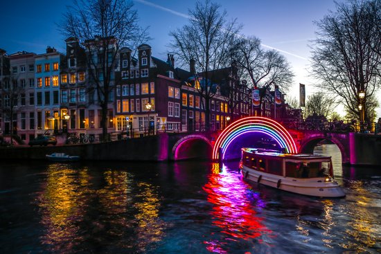 Beleuchtete Brücke mit buntem Neon-Regenbogen über einem Kanal in Amsterdam bei Dämmerung, Boot auf dem Wasser