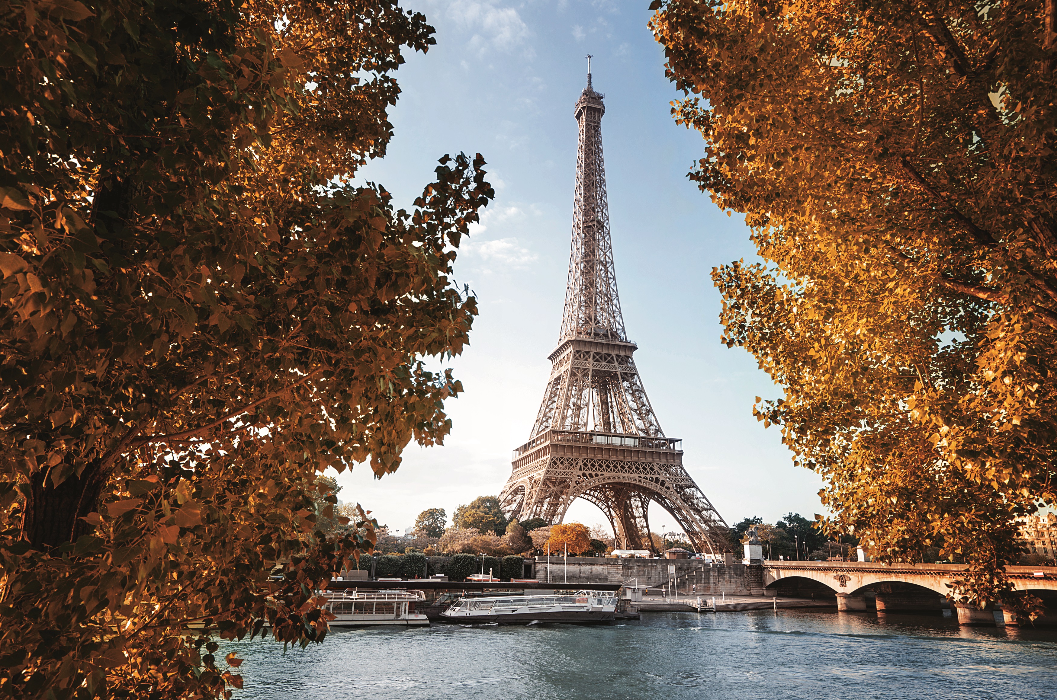 Eiffel Tower in Paris in autumn, surrounded by trees with orange leaves, river and bridge in the foreground.