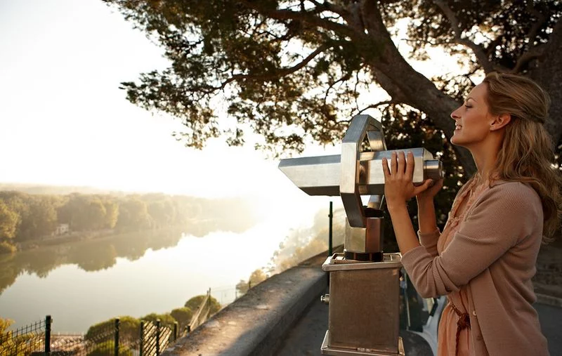A woman stands at a viewpoint in front of a public telescope, overlooking the river landscape at sunset, surrounded by green trees.