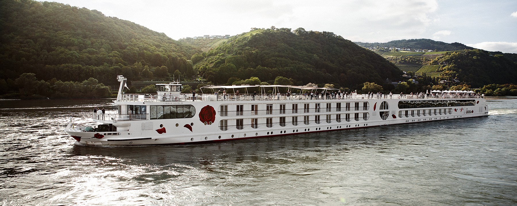 The A-ROSA BRAVA cruises through a green river landscape with wooded hills and vineyards.